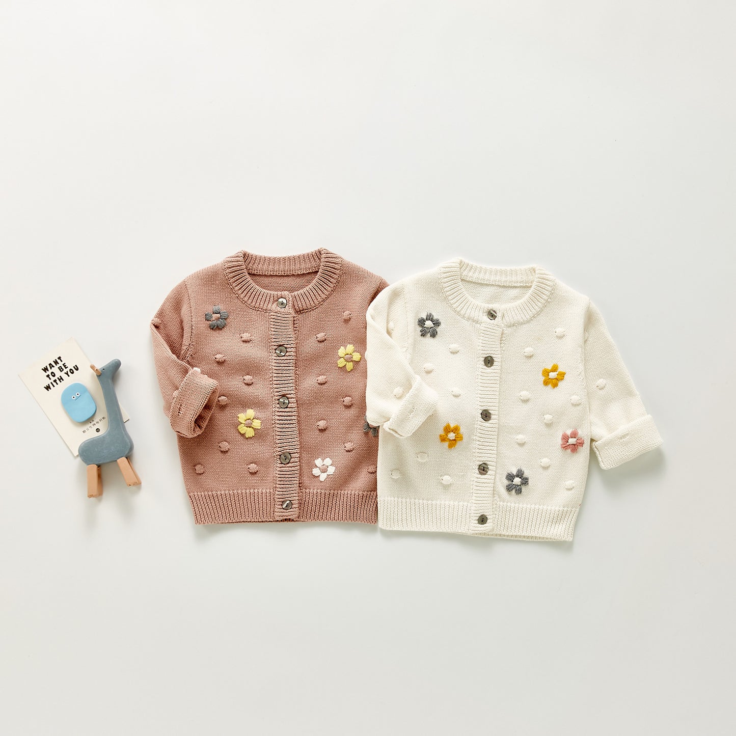 Two children's cardigans, one pink and one white, with floral patterns on a light gray background.