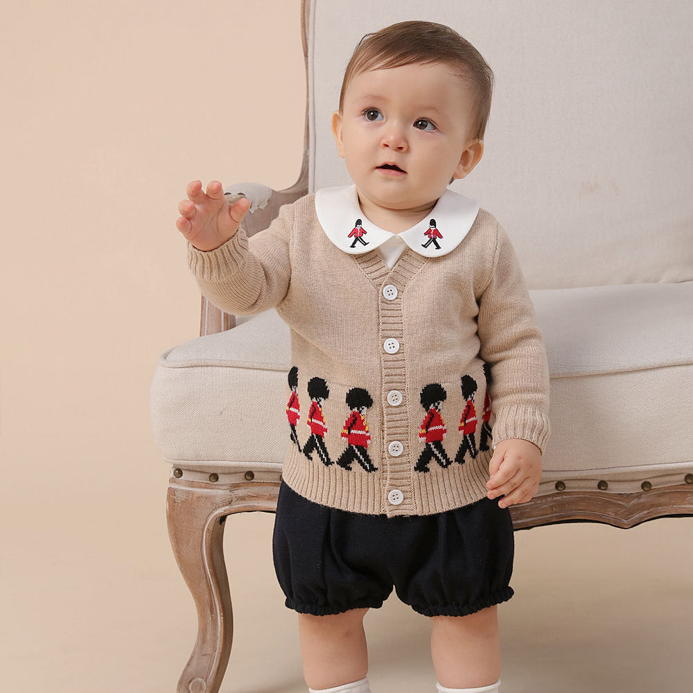 Baby wearing a beige cardigan with embroidered design and black shorts, standing in front of a wooden chair.