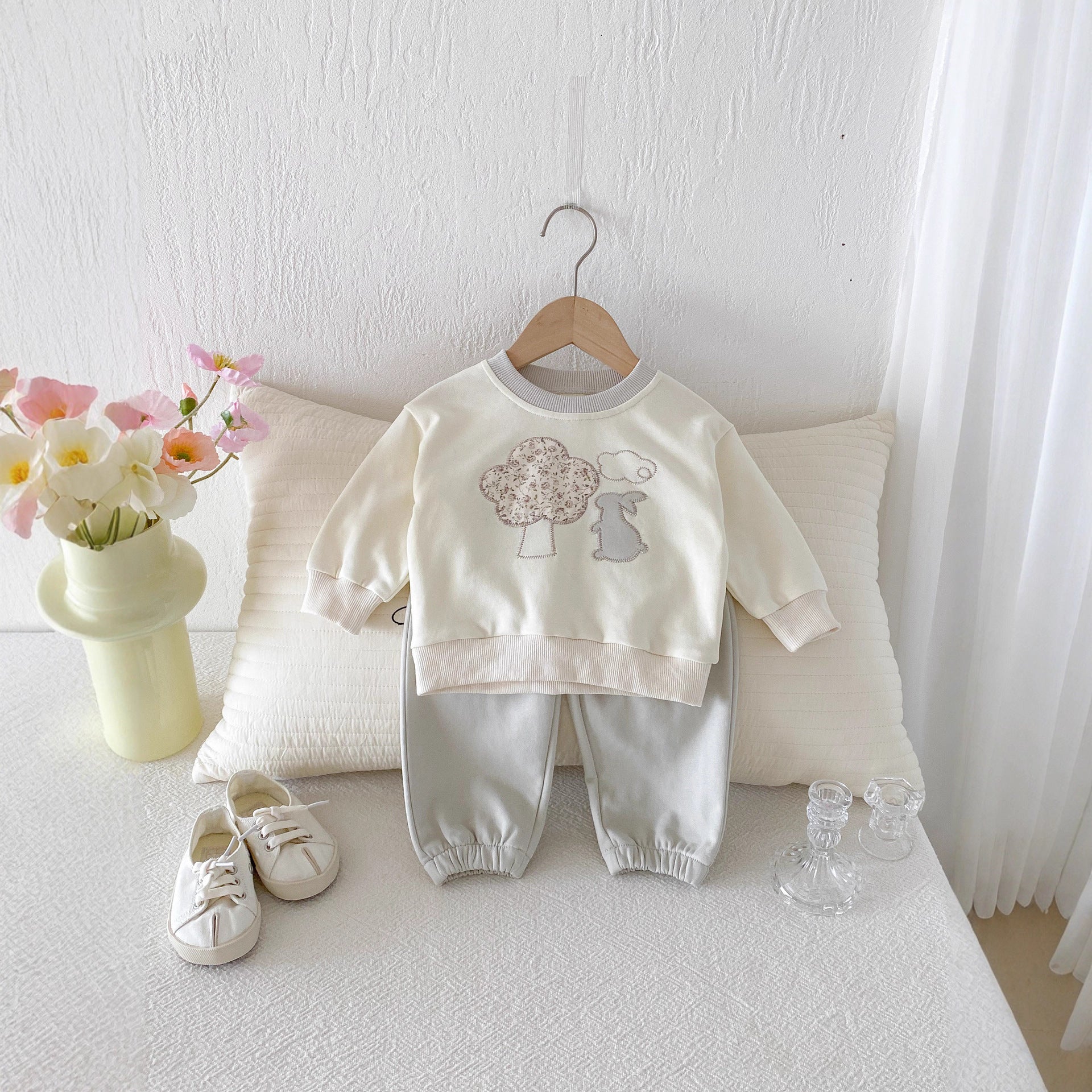 Baby outfit with sweatshirt and pants on a hanger, next to shoes and a vase with flowers on a light surface.
