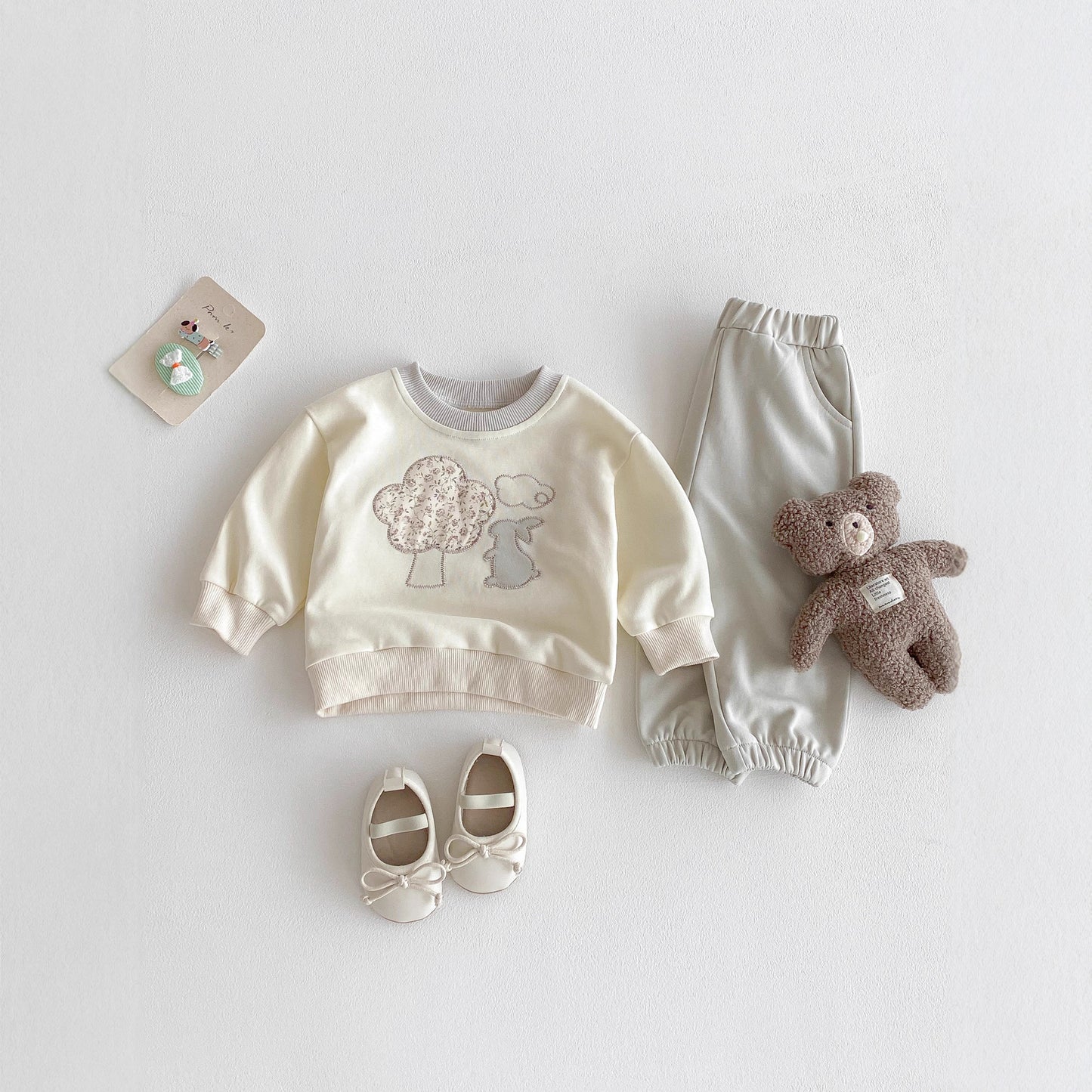 Baby outfit set with sweatshirt, pants, shoes, and teddy bear on a light gray background