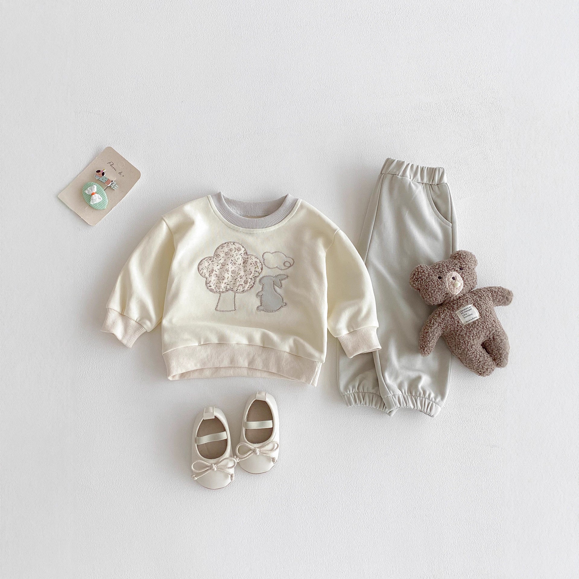 Baby outfit set with sweatshirt, pants, shoes, and teddy bear on a light gray background